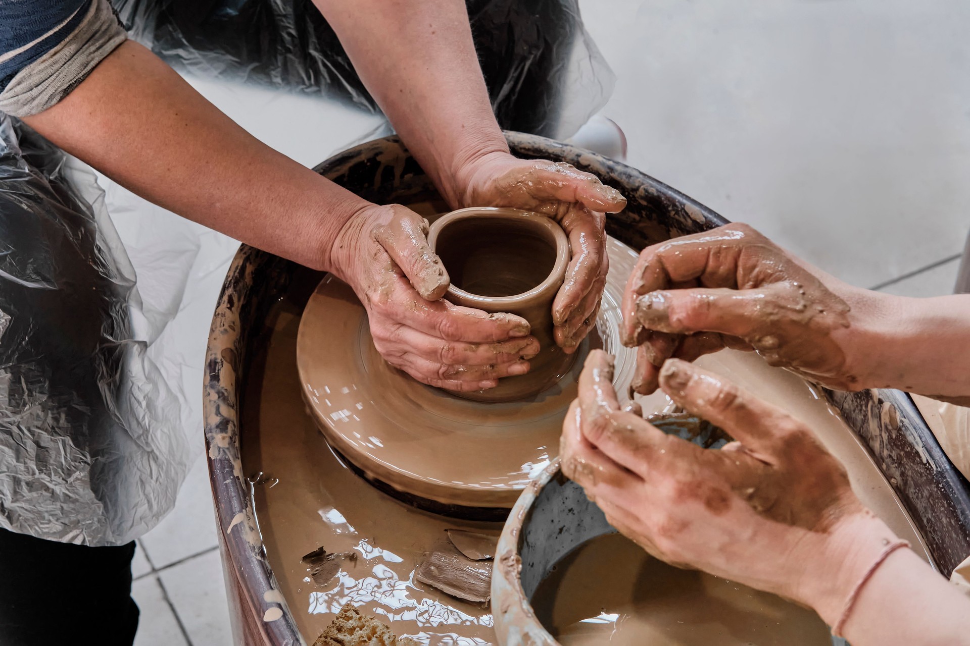Hands of senior woman and girl sculpting clay vase on potter's wheel at pottery training lesson.